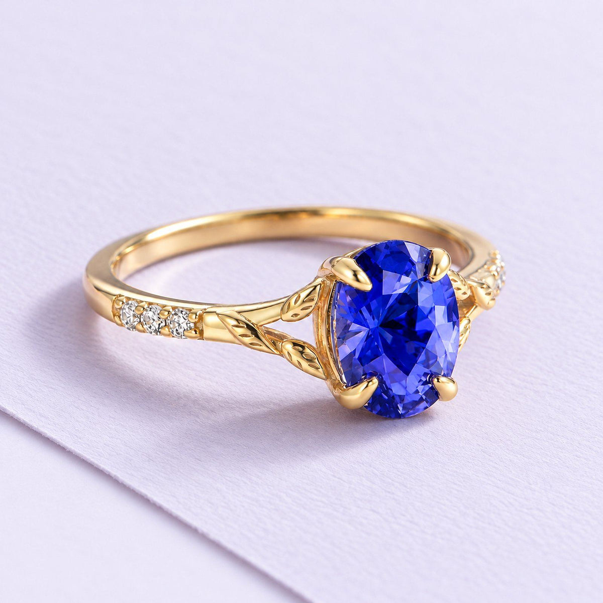 Yellow-gold blue sapphire leaf ring with clear shoulder accents, resting naturally on a broad sheet of pale lilac paper.