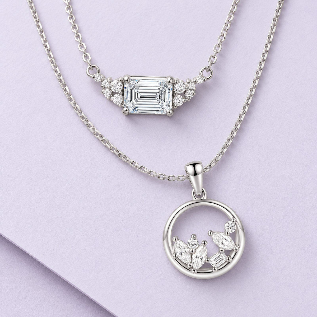 Two sterling-silver moissanite necklaces arranged in graceful layers on pale lilac paper, with emerald and circle pendants.