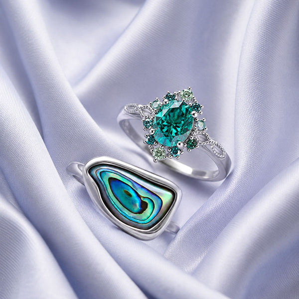 Sterling silver teal gemstone halo ring beside an abalone shell statement ring on lilac satin, Aquamarise collection banner