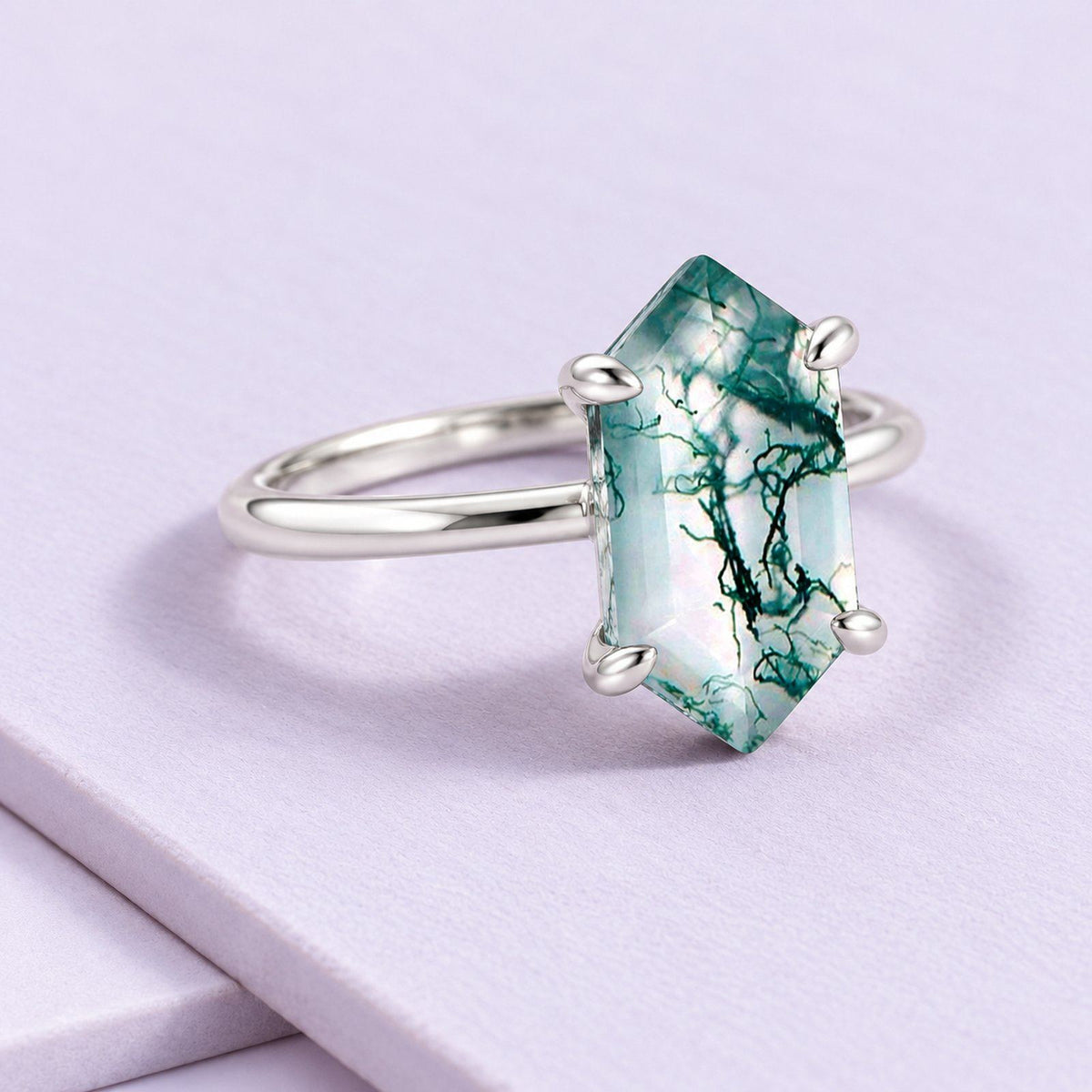 Elongated hexagon moss agate solitaire in silver, resting on full-size pale lilac tiles with a softly cropped diagonal join.