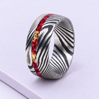 Damascus steel wedding band with red garnet and gold leaf inlay, resting on matte lilac tiles in soft daylight.