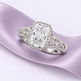 Radiant-cut moissanite ring with silver scrollwork resting on a continuous dusty lavender silk ribbon in soft daylight.