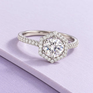 White-gold ring with a round lab-grown diamond, hexagonal halo and pavé shoulders, resting on broad pale-lilac tiles.