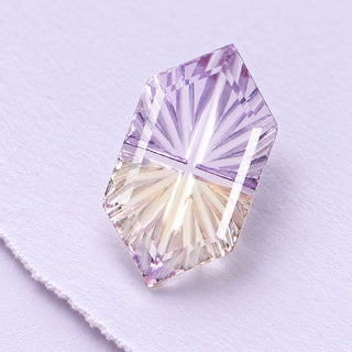 A pale ametrine Faelight gemstone shows its long hexagonal shape and precise fan-cut facets on softly textured lilac paper.