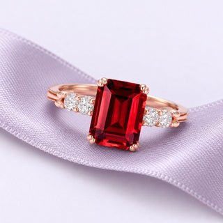 Emerald-cut red garnet ring in rose gold with clear shoulder accents, resting on a broad flowing lavender silk ribbon.