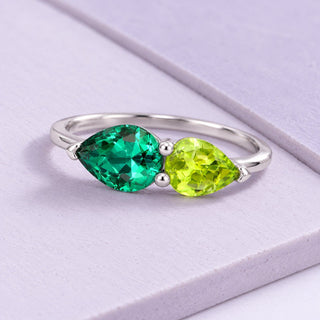 White-gold two-stone ring with an emerald and a yellow-green peridot in pear shapes, resting on broad pale-lilac tiles.