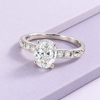 Oval lab-grown diamond ring with open white-gold filigree and clear side stones, angled naturally across pale-lilac tiles.