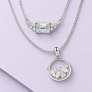 Two sterling-silver moissanite necklaces arranged in graceful layers on pale lilac paper, with emerald and circle pendants.