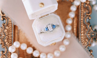 White emerald-cut center stone with pear-cut blue side stones in an open cream velvet ring box, on an ornate gilt tray beside a pearl strand
