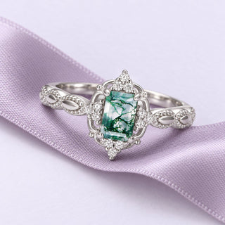 White-gold moss agate ring with a rectangular center and ornate openwork shoulders, resting on a long lavender ribbon.