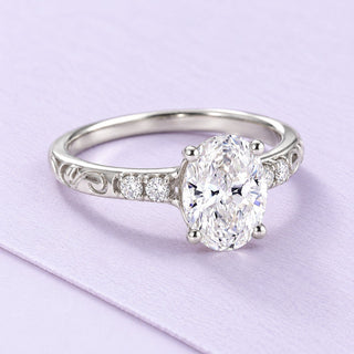 White-gold oval diamond ring with delicate filigree shoulders resting at a three-quarter angle on a large lilac paper sheet.