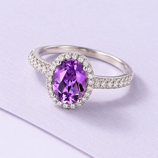 Oval purple amethyst ring with a clear halo and polished silver pavé band, photographed on a broad lilac paper sheet.