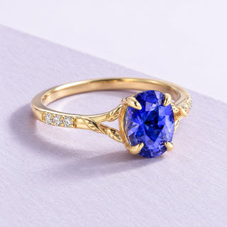 Yellow-gold blue sapphire leaf engagement ring with clear accents, photographed at an angle on textured pale lilac paper.