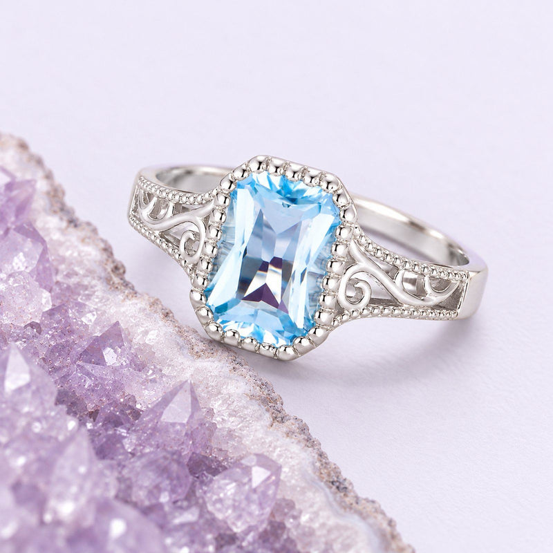 A sky-blue topaz ring with white-gold scrollwork rests beside a cropped lavender geode, showing its clear geometric facets.
