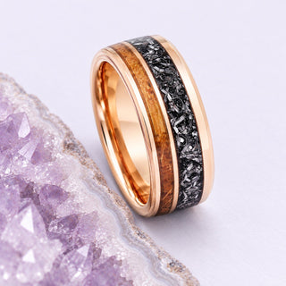 A rose-gold tungsten men's band with whiskey-barrel wood and dark meteorite inlays rests beside a cropped lavender geode.