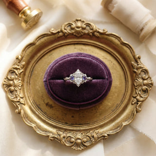 Vintage-style marquise diamond engagement ring with purple alexandrite side stones and an ornate white gold gallery, displayed in a purple velvet ring box on a gilded tray