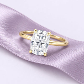 An elongated cushion diamond solitaire in yellow gold rests on a long lavender silk ribbon in a calm proposal still life.