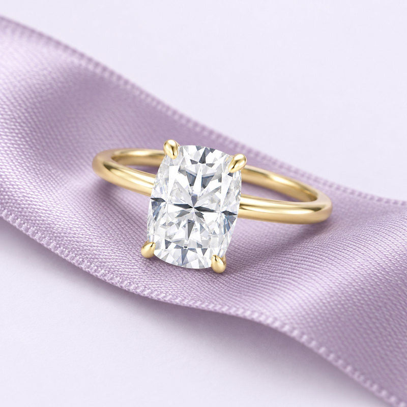 An elongated cushion diamond solitaire in yellow gold rests on a long lavender silk ribbon in a calm proposal still life.