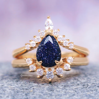 A teardrop-shaped blue sandstone gemstone set in a 14K solid yellow gold ring, with smaller clear stones around it, presented on a gray surface.