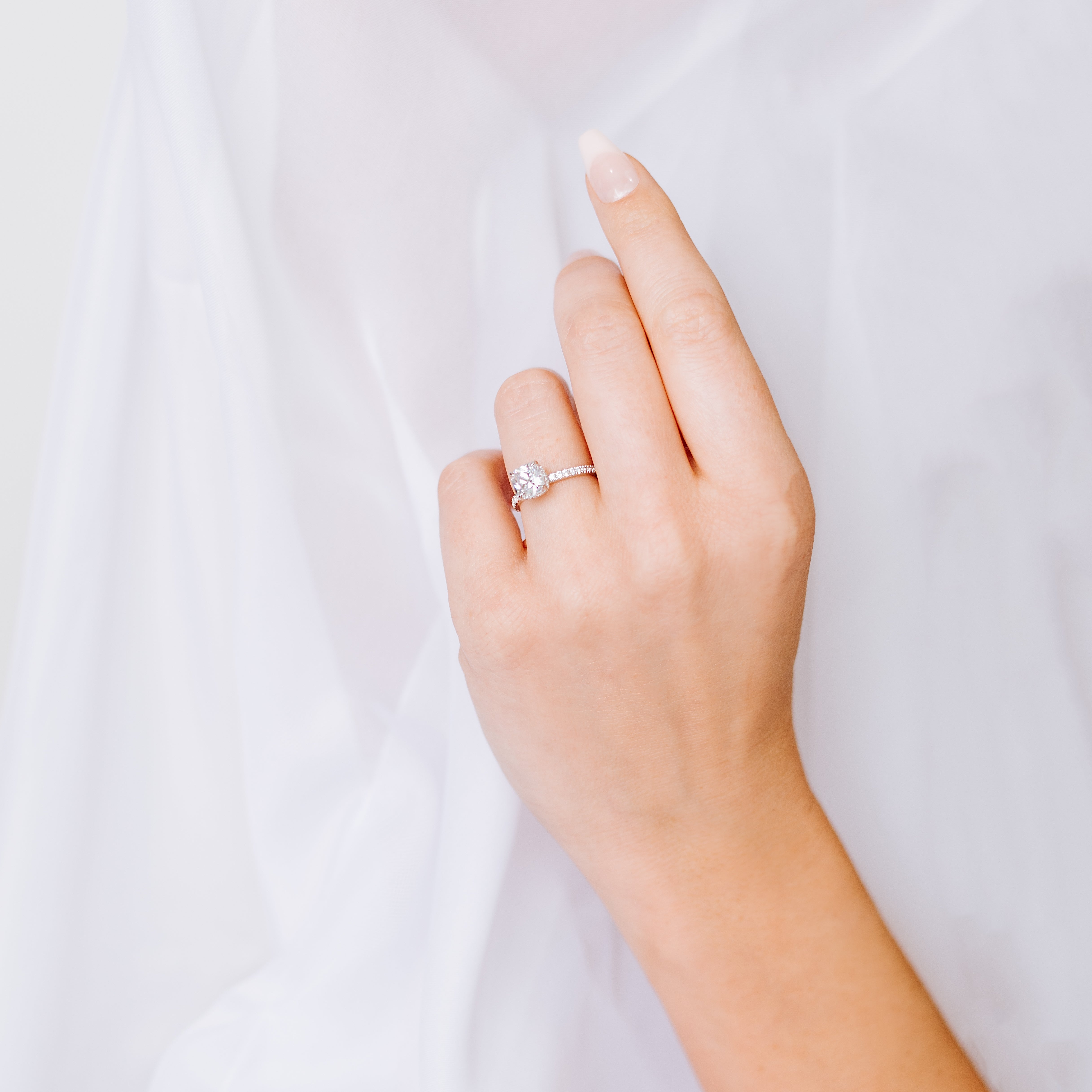 Three carat square cushion cut moissanite engagement ring with a hidden halo and a thin pave band, worn on a hand against white fabric