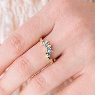 Yellow gold wedding band with crown design and blue moissanite and colorless moissanite accents on hand over light background