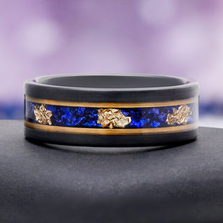 Blue Sapphire & Gold Leaf Brushed Men's Wedding Band in Black Tungsten (8mm) by Aquamarise, resting on a grey stone to show the sapphire and gold leaf inlay.