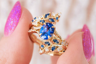 Hand holding gold ring with leaf design and blue sapphire center stone 