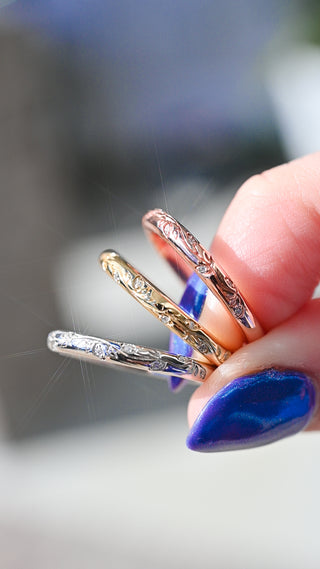 Hand holding three slim engraved rings in silver, gold, and rose gold tones, with sunlight reflecting off the metallic surfaces and a blue-painted thumbnail visible.