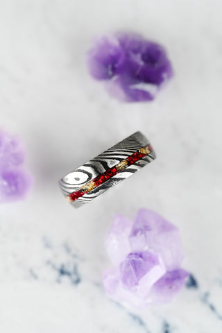 Silver ring with red gemstone on a white surface with purple flowers. Damascus steel men's wedding bands