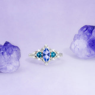 Silver ring with blue gemstone surrounded by purple crystals on a light purple background