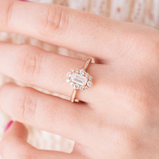 Rose gold engagement ring with emerald cut diamond center stone and accents on hand over a light background 