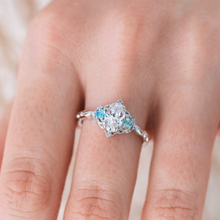 White gold engagement ring with oval cut diamond center stone and aquamarine accents on hand over a light background 