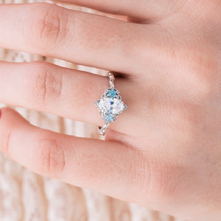 White gold engagement ring with oval cut diamond center stone and aquamarine accents on hand over a light background 