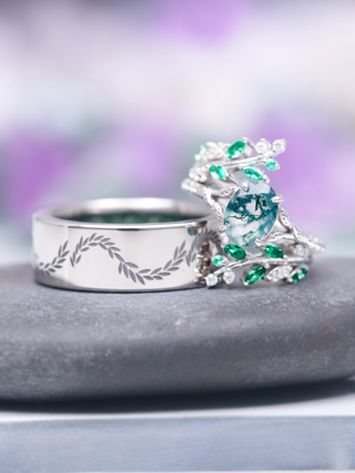 Silver ring with engraved design and green gemstones on a gray surface with a blurred purple background