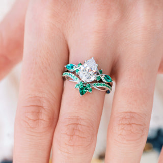White gold engagement ring set with teardrop cut diamond center stone and lab-grown emerald accents on hand over a light background 