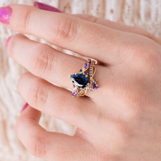 Yellow gold engagement ring set with teardrop cut blue sandstone center stone and amethyst accents on hand over a light background 