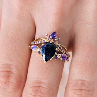 Yellow gold engagement ring set with teardrop cut blue sandstone center stone and amethyst accents on hand over a light background 