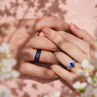 Couple's clasped hands wearing the Galaxy Orion Nebula and Blue Sapphire Couples Ring Set by Aquamarise, a black band with blue inlay beside a pear blue sapphire ring