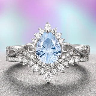 An elegant two-piece engagement ring set featuring a teardrop-shaped natural aquamarine centerpiece with fantasy cut gemstones, surrounded by white gold and Moissanite accent stones on a twisted band, with a matching wedding band in the background.