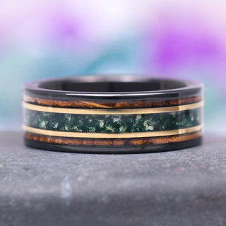 An 8MM two-tone gold and black tungsten wedding band with an inlay of whiskey barrel wood and moss agate stone, displayed against a gradient purple background.