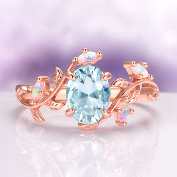 Aquamarine & Fire Opal Leaf Engagement Ring in 14K Solid Rose Gold by Aquamarise Gold, featuring an oval aquamarine center with white fire opal accents on a rose gold leaf-inspired band.