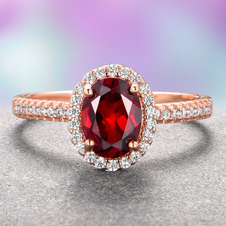 An oval red garnet ring with a halo of smaller accent stones set in a 14K solid rose gold band.