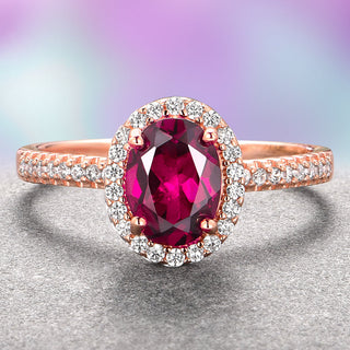 An oval ruby engagement ring with a halo of smaller diamonds set in a rose gold band, displayed against a blurred multicolored background.