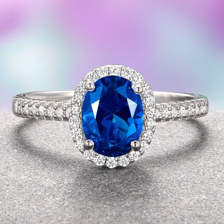 An oval blue sapphire engagement ring with a halo of white gold and diamond accents on a purple and gray background.