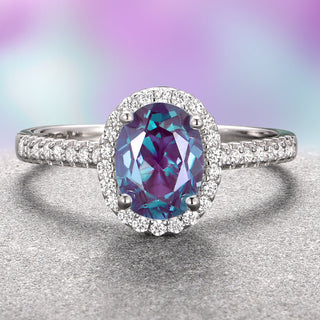 An oval-shaped Alexandrite gemstone engagement ring with a halo of either moissanite or diamond accents set in a 14K white gold band, displayed against a gradient purple and pink background.