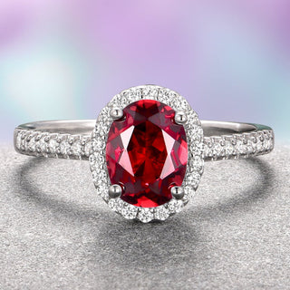 An oval red garnet engagement ring with a halo of white gold and accent stones set on a textured metal band.
