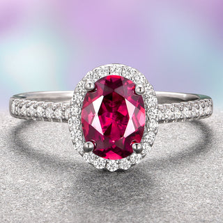 An oval ruby engagement ring with a halo of smaller diamonds set in a 14K white gold band, displayed against a gradient purple and gray background.