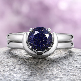 A sterling silver engagement ring set featuring a round-cut blue goldstone center stone with a crescent moon-inspired band, displayed against a purple background.