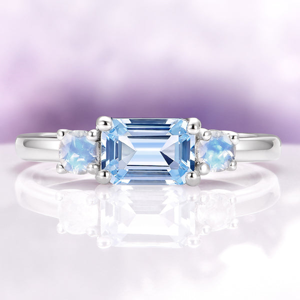 Emerald Cut Aquamarine and Moonstone Engagement Ring in 14K White Gold by Aquamarise Gold, featuring a luminous aquamarine center flanked by glowing moonstones in a sleek white gold setting.
