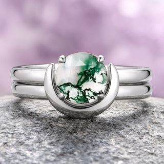 A sterling silver ring set featuring a round-cut moss agate center stone with a crescent moon-inspired band, displayed on a rock.